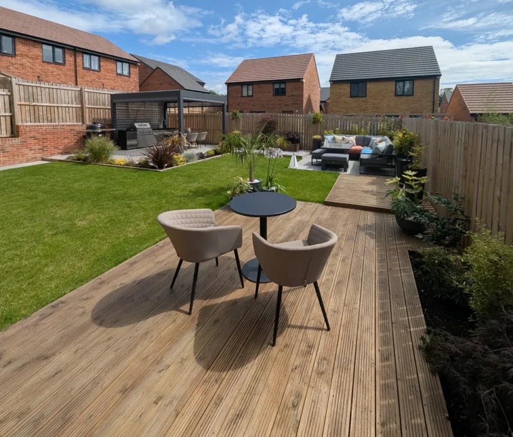 Premier Gardens A modern garden with a wooden decking, two beige chairs and a round table, green lawn, soft landscaping, outdoor seating area, plants, and houses in the background under a partly cloudy sky. Landscaping Newcastle