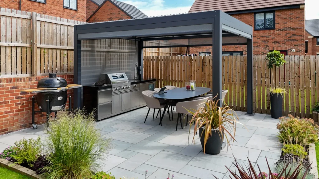 Premier Gardens A modern back garden by Premier Gardens features a covered outdoor kitchen with a stainless steel grill, round dining table, lush soft landscaping, and potted greenery on a tiled patio. Wooden fencing and brick houses provide the perfect backdrop. Landscaping Newcastle
