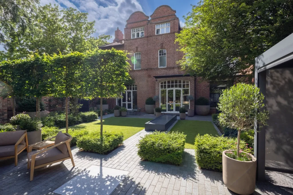 Premier Gardens A well-maintained courtyard garden by Premier Gardens features modern outdoor seating, manicured hedges, potted plants, and impressive hard landscaping around a brick house with large windows and arched roof details under a partly cloudy sky. Landscaping Newcastle