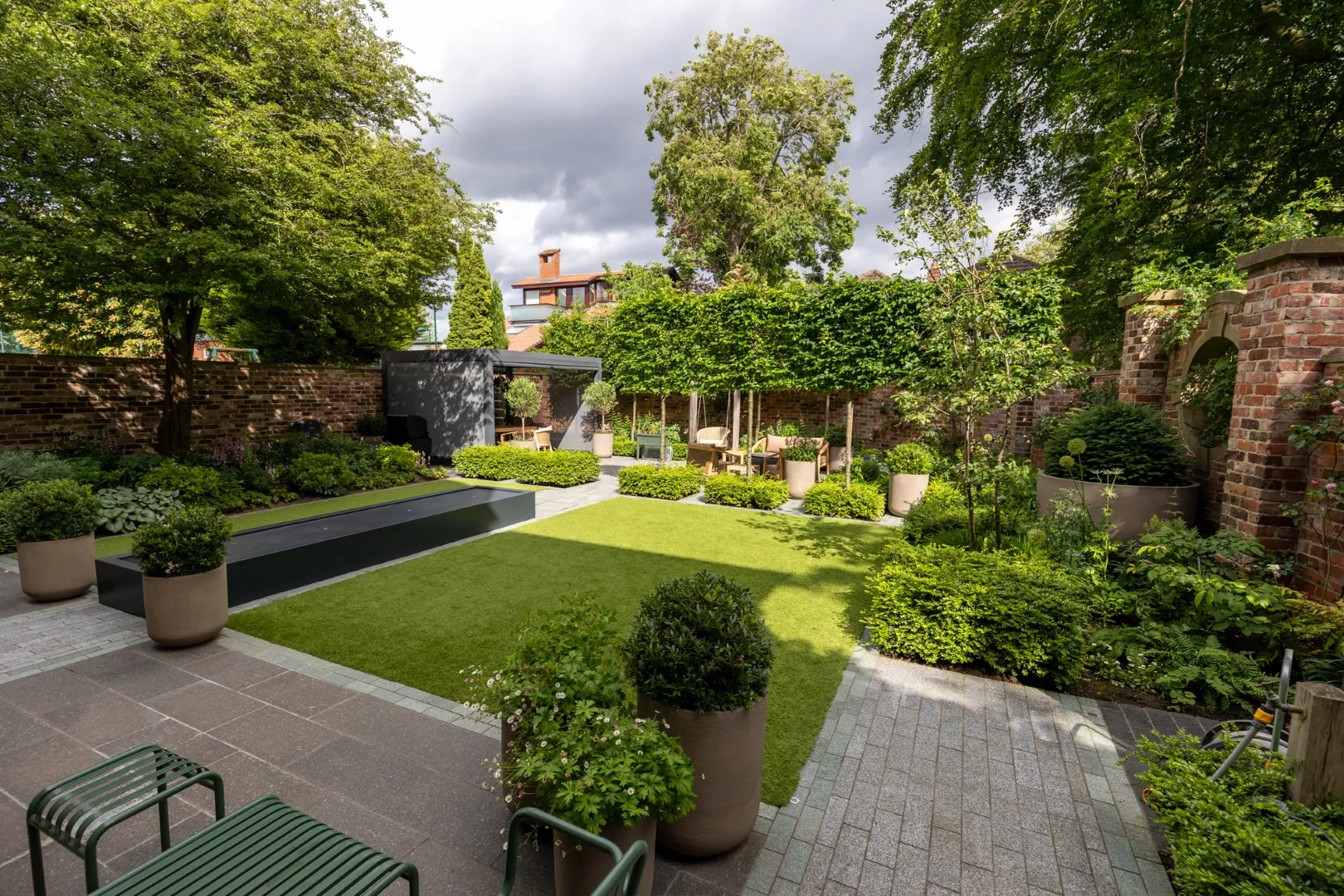 Premier Gardens A modern garden by Premier Gardens featuring soft landscaping with neatly trimmed grass, potted plants, green shrubs, and tall trees. Paved walkways, patio installation North East, outdoor seating, and a rectangular water feature are enclosed by brick walls. Landscaping Newcastle