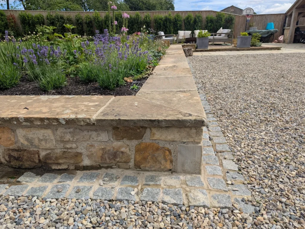 Premier Gardens A stone garden wall borders a raised flower bed with purple and pink blooms, showcasing expert garden design Northumberland. Pebbles and grey paving stones form a path, while patio furniture and a basketball hoop add charm in the background. Landscaping Newcastle